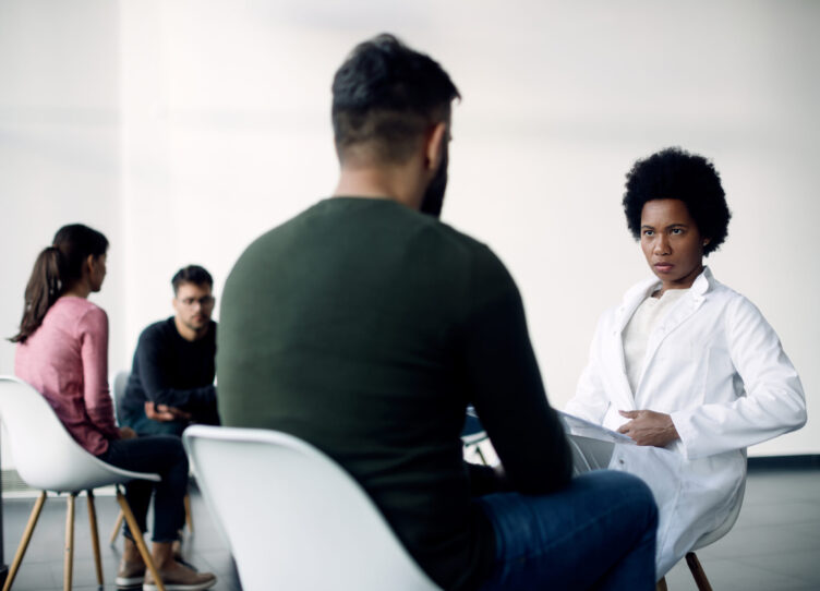 Black female doctor communicating with a man during group psychotherapy meeting.