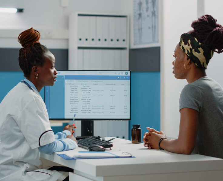African american doctor and woman looking at computer screen for information about appointment and healthcare system. Patient sitting at desk with medic for annual checkup visit.